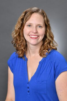 This is a headshot photo with a gray background. There is a white woman with blue eyes and wavy, dark blonde, shoulder length hair who is wearing a royal blue, v-neck shirt. She has a smile showing her teeth and light red lipstick. 