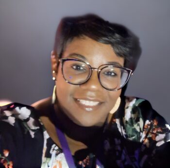 Headshot photo of a Black woman with short hair that has a wave across one side of her forehead. She is wearing large, mostly round, dark purple glasses with a beautiful gold accent at the corner. She has several earrings and is wearing a black blouse with a floral print. 